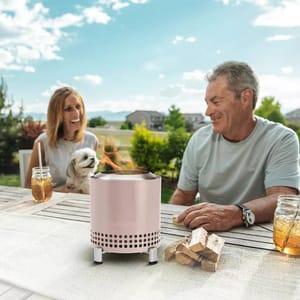 A portable outdoor fire pit and a pair of wooden logs on a patio table with two smiling people and a dog in the background.