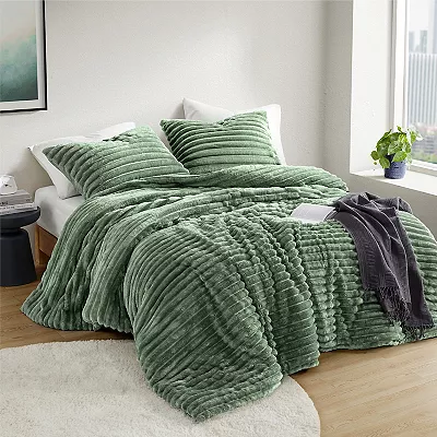 A green ribbed comforter set with matching shams and a decorative grey throw blanket on a bed, set against a neutral room decor.