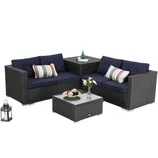 Outdoor sectional furniture set with dark wicker frame, navy cushions, and striped pillows includes a corner table with a glass top and a matching square coffee table. A decorative lantern and floral vase are on the tables.