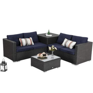 Outdoor sectional furniture set with dark wicker frame, navy cushions, and striped pillows includes a corner table with a glass top and a matching square coffee table. A decorative lantern and floral vase are on the tables.
