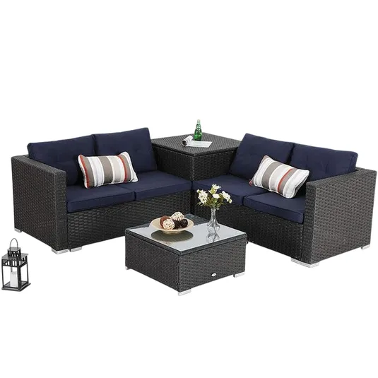 Outdoor sectional furniture set with dark wicker frame, navy cushions, and striped pillows includes a corner table with a glass top and a matching square coffee table. A decorative lantern and floral vase are on the tables.