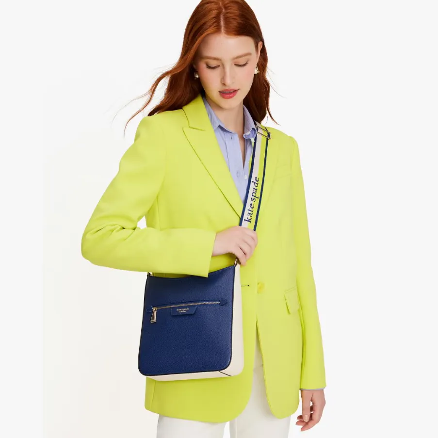 A woman is wearing a bright lime blazer, blue button-up shirt, white pants, and has a blue and cream crossbody bag.
