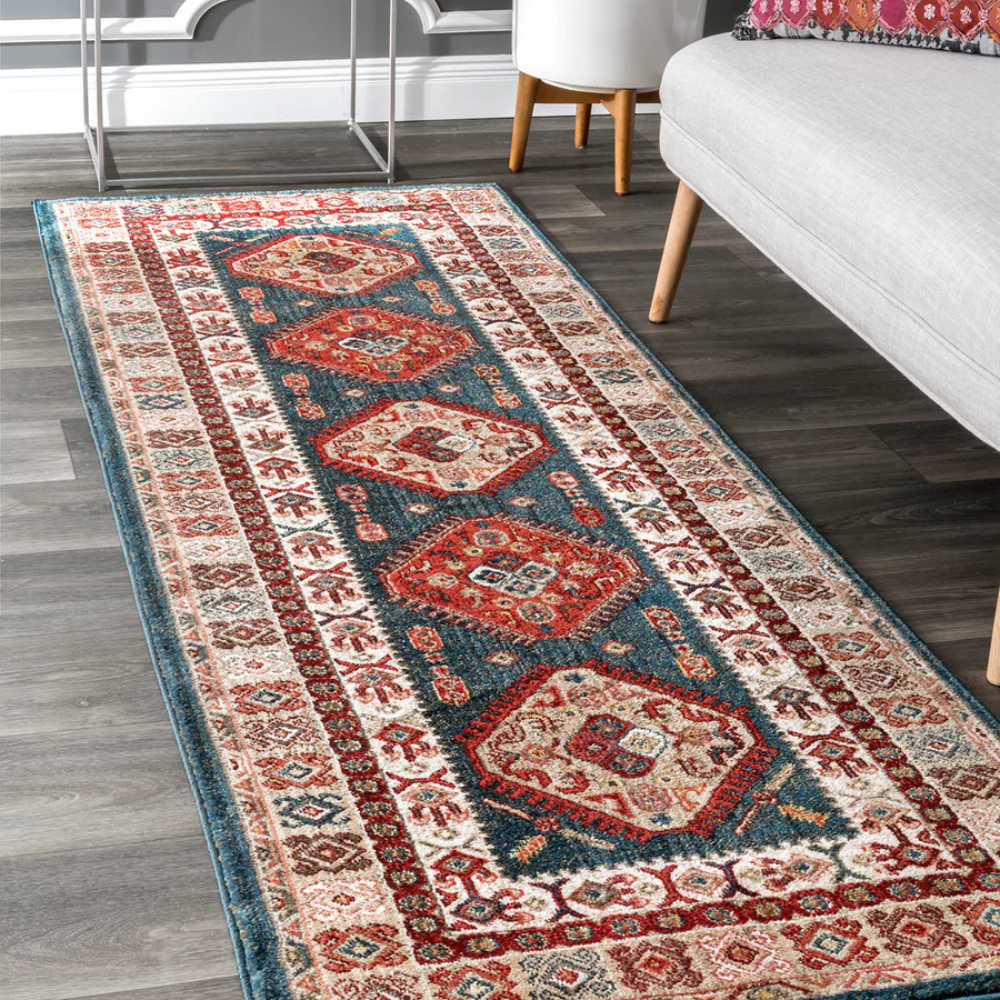 A decorative area rug with a traditional Persian-style pattern featuring blue, red, and cream colors, placed on a dark wood floor next to a gray sofa and white furniture.