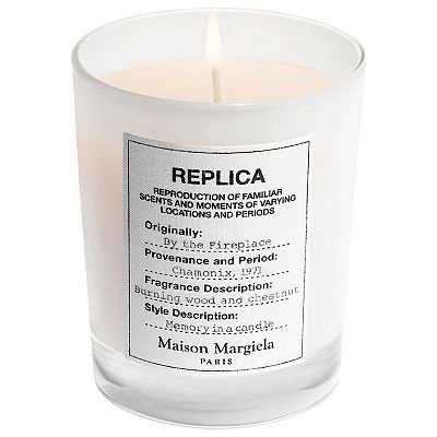 A scented candle by Maison Margiela with a label describing \“By the Fireplace\“ fragrance, indicating woody, chestnut scent characteristics.