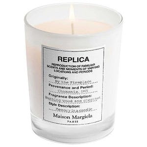 A scented candle by Maison Margiela with a label describing \“By the Fireplace\“ fragrance, indicating woody, chestnut scent characteristics.
