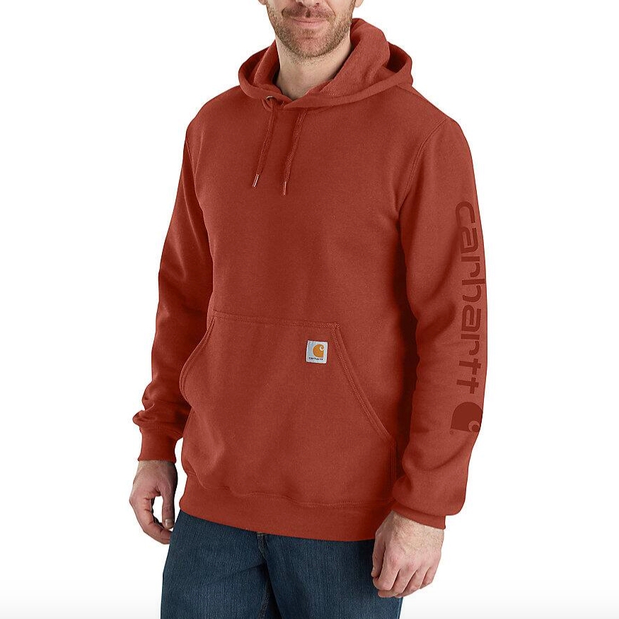 A red Carhartt hoodie with a front pocket, drawstring hood, and Carhartt logo on the sleeve. It is paired with dark jeans.