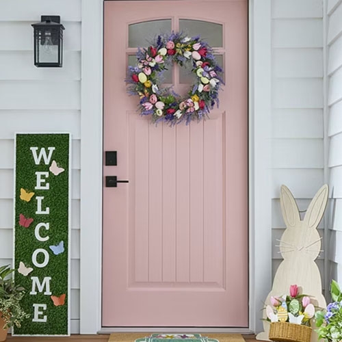 A colorful wreath with pastel flowers and Easter eggs decorates a pink door, complemented by a bunny cutout, a \“Welcome\“ sign with butterfly accents, and a small potted plant on a porch.