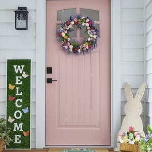 A colorful wreath with pastel flowers and Easter eggs decorates a pink door, complemented by a bunny cutout, a \“Welcome\“ sign with butterfly accents, and a small potted plant on a porch.
