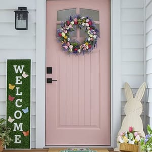 A colorful wreath with pastel flowers and Easter eggs decorates a pink door, complemented by a bunny cutout, a \“Welcome\“ sign with butterfly accents, and a small potted plant on a porch.