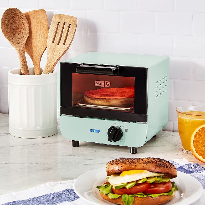 A turquoise Dash Mini Toaster Oven with a bagel inside is shown on a kitchen countertop next to wooden utensils and a sandwich on a plate.