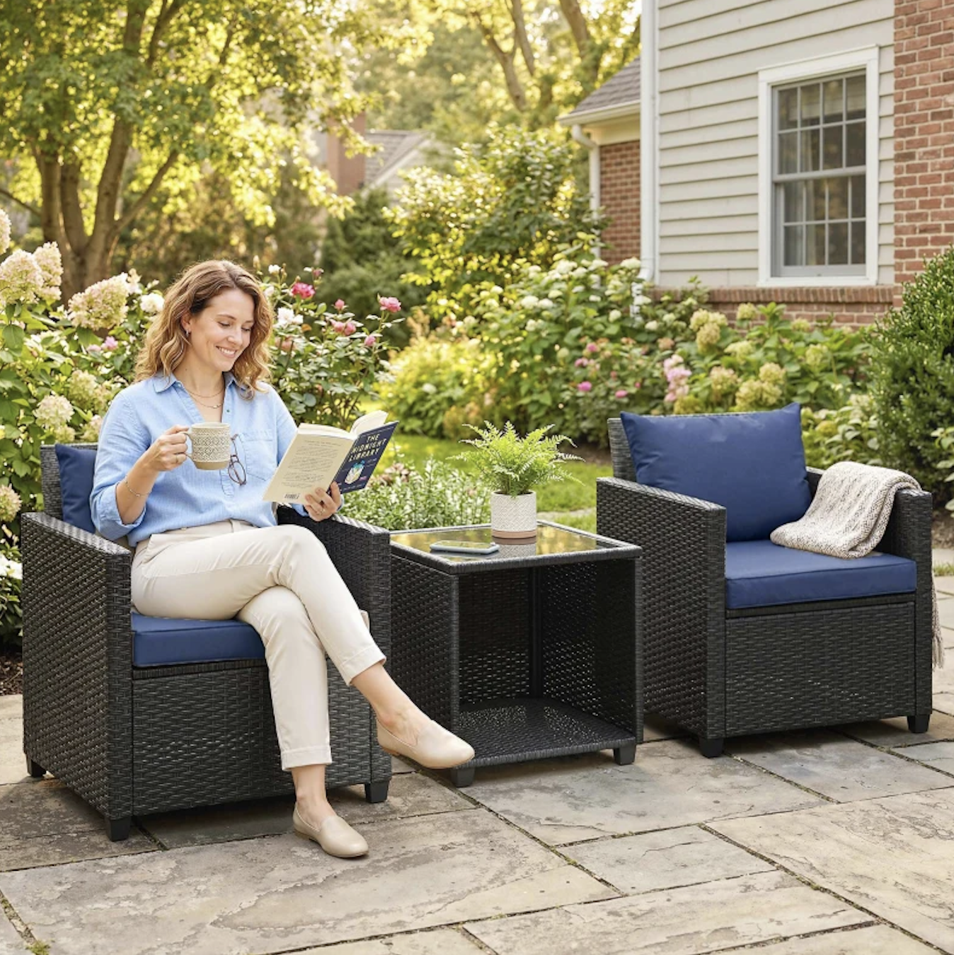 Outdoor patio conversation set featuring two wicker chairs with blue cushions, a side table with a glass top, and a woman enjoying a book and drink.