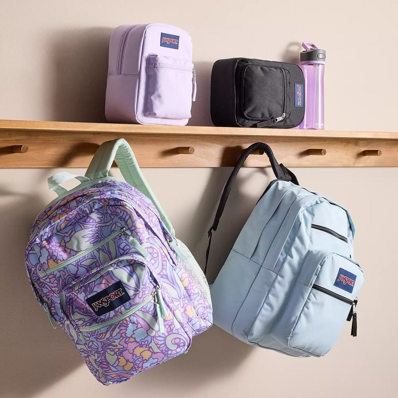 Four backpacks displayed on pegs: a lavender pouch, a black pouch with water bottle, a purple patterned backpack, and a light blue backpack.