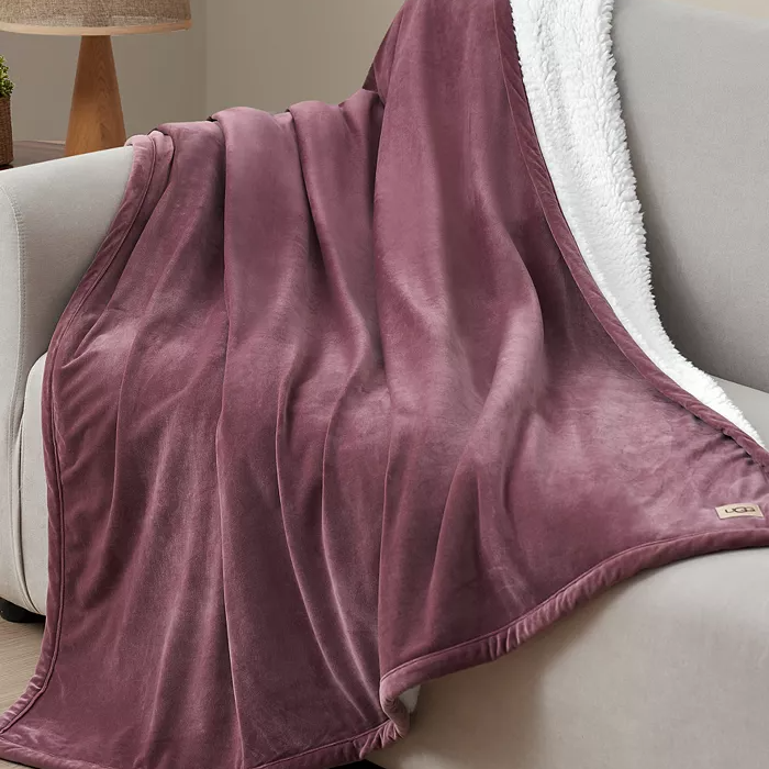 UGG Dasha Plush Throw features a soft plum-colored exterior and a cozy white sherpa lining, draped over a light-colored couch.
