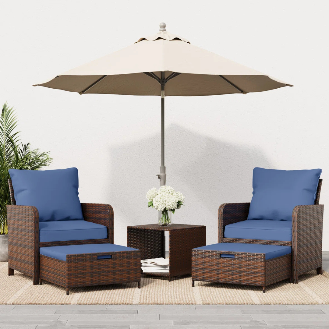 Outdoor patio set featuring two wicker chairs with blue cushions, matching ottomans, a small wicker table, a beige umbrella, and a vase of white flowers.