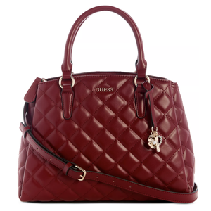 A red quilted handbag with two top handles, a detachable shoulder strap, and decorative charms. It features the GUESS logo on the front.