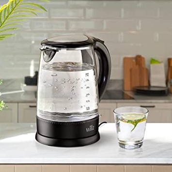 A transparent glass electric kettle with a black plastic handle and base is shown next to a glass with a lemon slice on a kitchen counter.