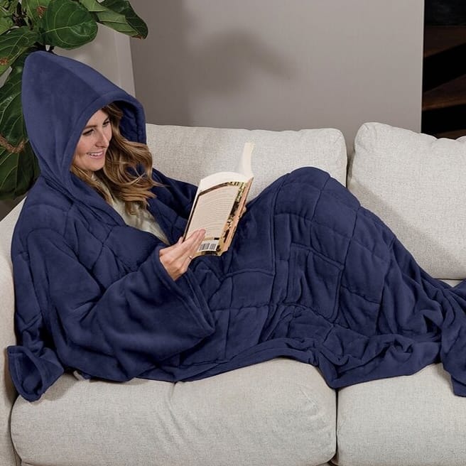 A person is reclining on a couch, reading a book while wearing a cozy, navy blue hooded blanket with sleeves.