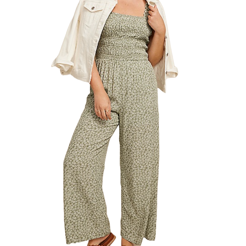 A model wears a green floral smocked jumpsuit with a white denim jacket draped over her shoulders, paired with brown sandals.