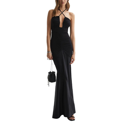 A woman is wearing a long black halter-neck evening gown with a cross-strap design and ruched detailing, paired with black sandals and holding a small black purse.