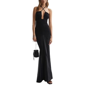 A woman is wearing a long black halter-neck evening gown with a cross-strap design and ruched detailing, paired with black sandals and holding a small black purse.