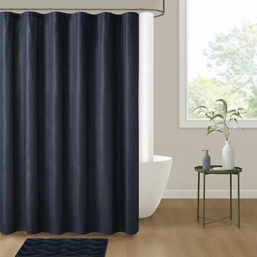 A navy blue shower curtain hangs in a bathroom with a freestanding tub. Nearby, a small round table holds a plant and two decorative bottles. A dark blue bath mat is placed on the wooden floor.