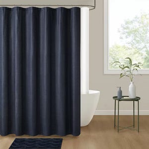 A navy blue shower curtain hangs in a bathroom with a freestanding tub. Nearby, a small round table holds a plant and two decorative bottles. A dark blue bath mat is placed on the wooden floor.