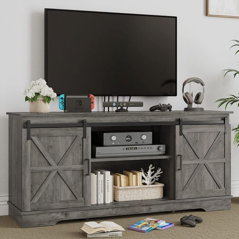 A wooden TV stand with barn-style sliding doors holds a flat-screen TV, game console, controller, headphones, and books. A plant and a flower pot are placed on top for decoration.