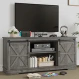 A wooden TV stand with barn-style sliding doors holds a flat-screen TV, game console, controller, headphones, and books. A plant and a flower pot are placed on top for decoration.