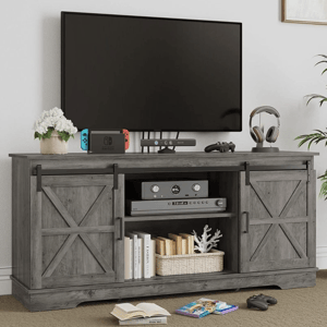 A wooden TV stand with barn-style sliding doors holds a flat-screen TV, game console, controller, headphones, and books. A plant and a flower pot are placed on top for decoration.