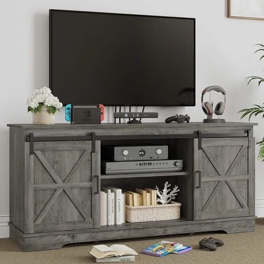 A wooden TV stand with barn-style sliding doors holds a flat-screen TV, game console, controller, headphones, and books. A plant and a flower pot are placed on top for decoration.