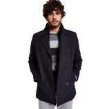 A man is wearing a dark double-breasted coat over a gray and black striped sweater, paired with light gray pants.