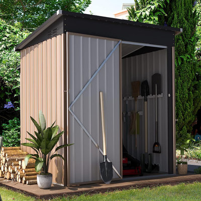 A compact, metal storage shed with brown and grey panels, a sloped roof, and double sliding doors, set in a garden with plants and stacked firewood.