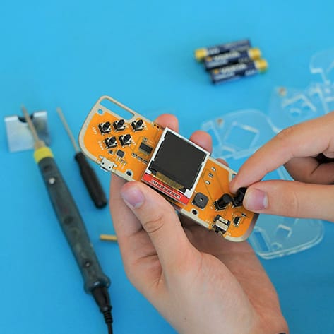Hands assembling an electronic circuit board shaped like a handheld gaming console with tools nearby.