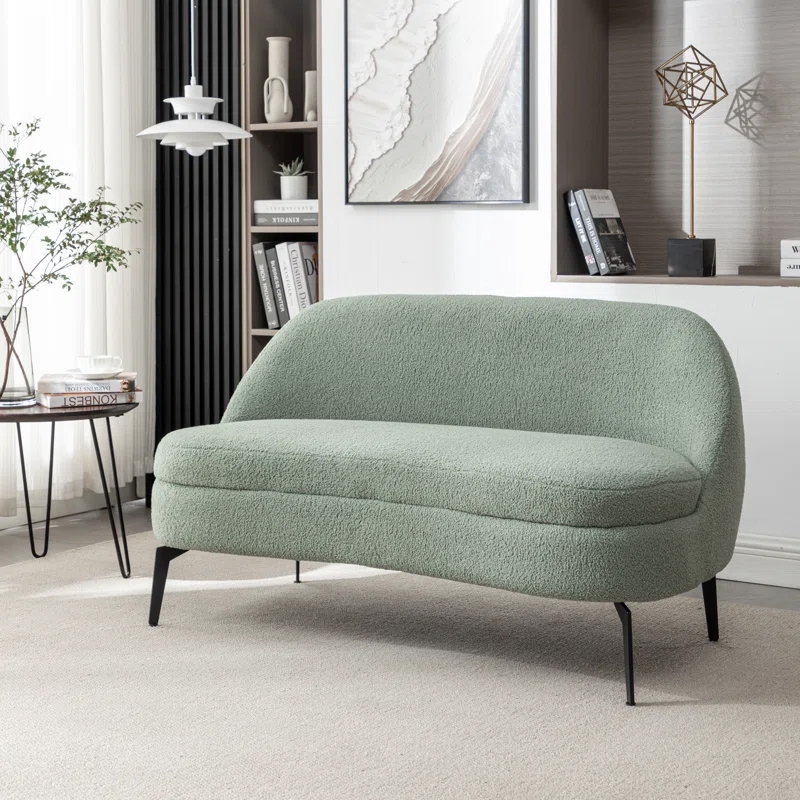 A light green loveseat with a curved backrest and plush texture, set on slim black metal legs. It's placed in a modern living room with neutral tones and minimalist decor elements.