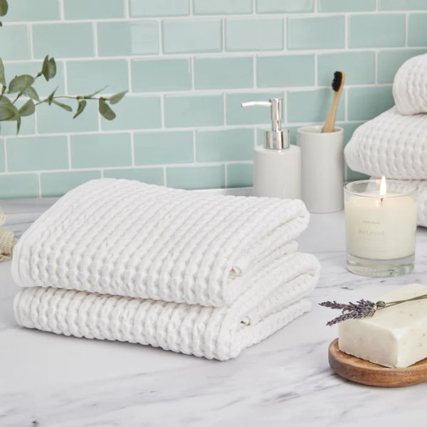 Two white waffle-patterned towels are neatly folded on a marble countertop, surrounded by a soap dispenser, a toothbrush in a holder, a lit candle, and a bar of soap. The background features a light green tiled wall.
