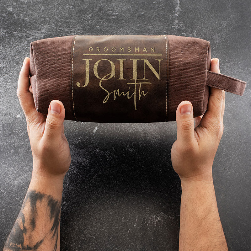 A brown toiletry bag with \“Groomsman John Smith\“ printed on it is held by two hands against a dark background.