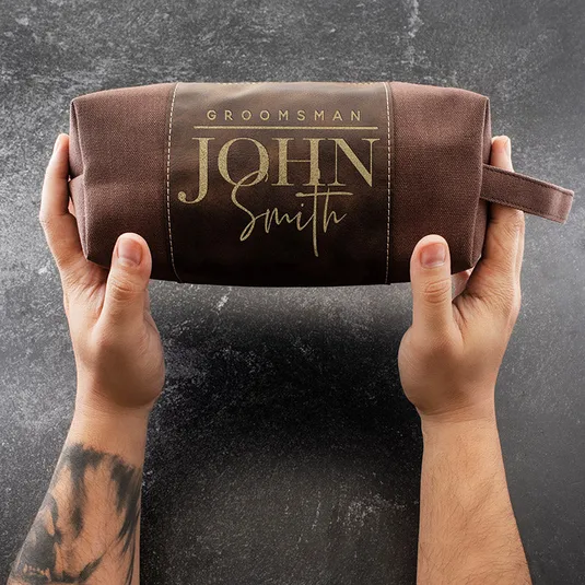 A brown toiletry bag with \“Groomsman John Smith\“ printed on it is held by two hands against a dark background.