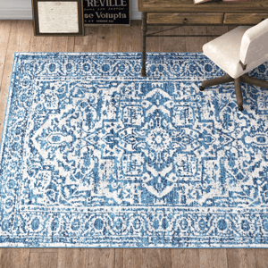 A blue and white patterned rug lies on a wooden floor, partially placed under a beige chair and a wooden table with decor.