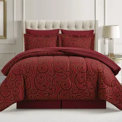 A red comforter set with gold swirl patterns, including matching pillow shams, is on a bed with a tufted headboard and accompanied by a bedside lamp.