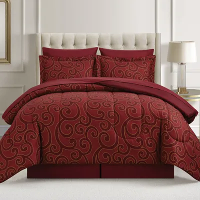 A red comforter set with gold swirl patterns, including matching pillow shams, is on a bed with a tufted headboard and accompanied by a bedside lamp.