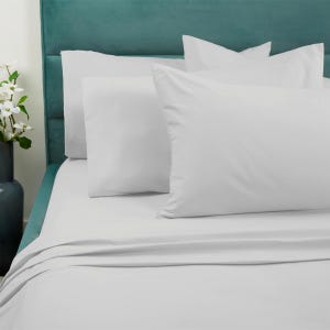 A 6-piece sheet set including white pillowcases and fitted sheets with a smooth texture, displayed on a bed with a teal headboard. A dark vase with white flowers is placed nearby.