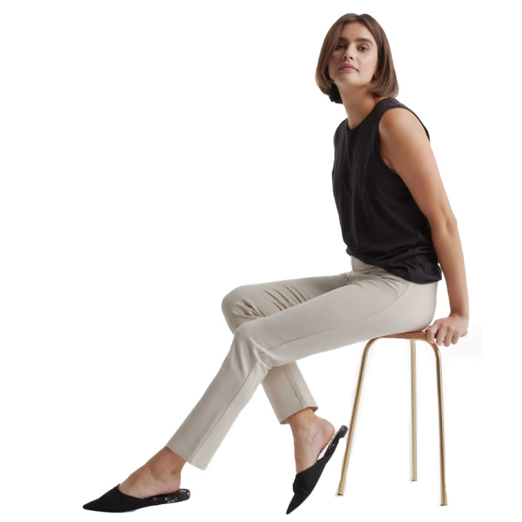 A woman wearing beige pants, a sleeveless black top, and black slip-on shoes sits on a stool.