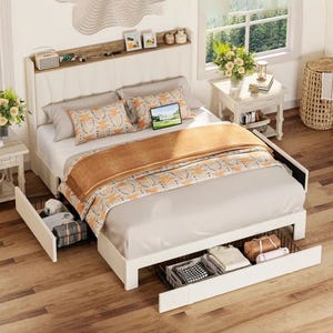 A white storage bed features drawers on both sides and at the foot of the bed for added storage. It includes a built-in charging station in the headboard, which also has a shelf displaying decorative items. The bedding is light with orange accents.