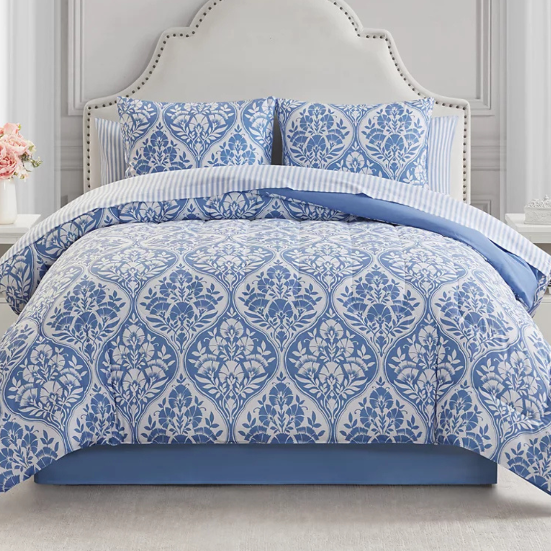 A bed with a blue and white floral patterned comforter and matching pillow shams, featuring a striped sheet underneath. The headboard is white with a studded border.