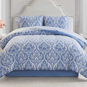 A bed with a blue and white floral patterned comforter and matching pillow shams, featuring a striped sheet underneath. The headboard is white with a studded border.