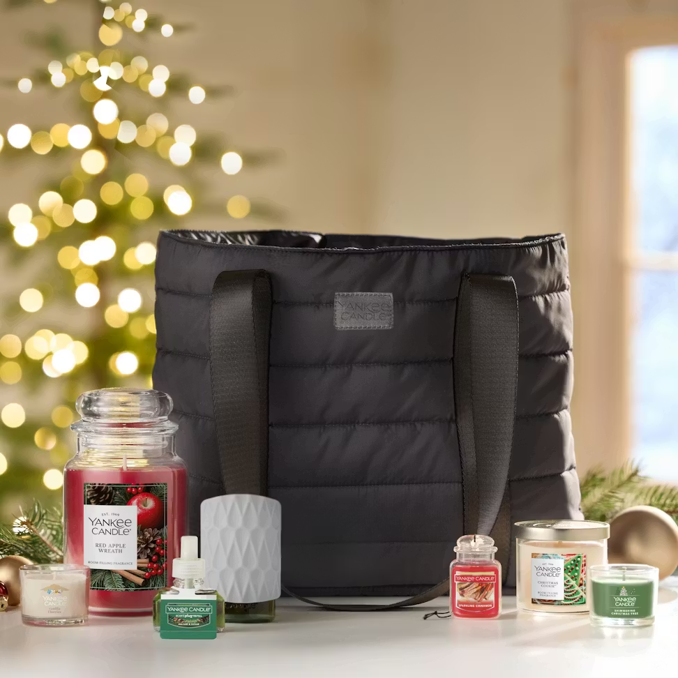 A black tote bag is surrounded by various Yankee Candle products: a jar candle (Red Apple Wreath), a smaller candle jar, a diffuser with oil, and a candle in a tin, against a festive holiday background.