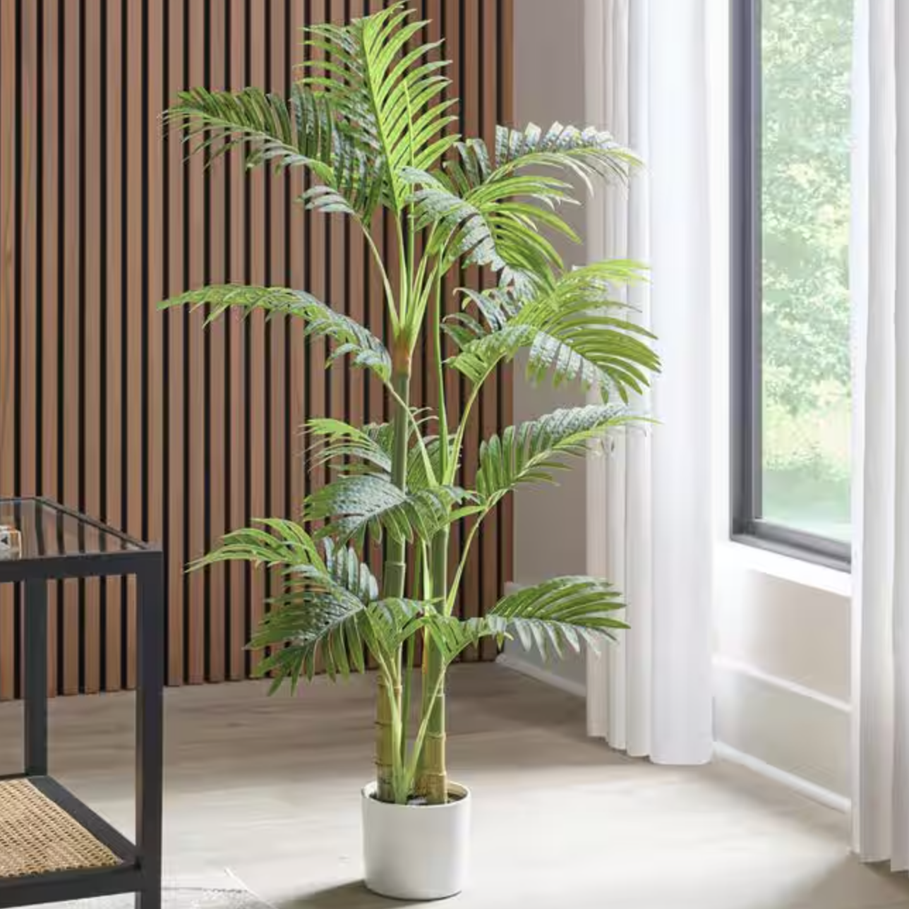 A 4' artificial palm tree with arching fronds in a white pot, placed in a modern interior space near a window with natural light, next to a wood-paneled wall.