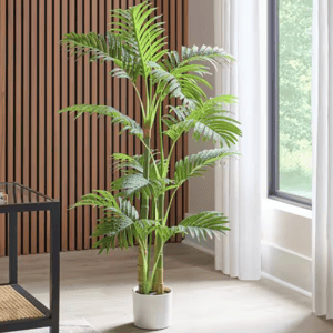 A 4' artificial palm tree with arching fronds in a white pot, placed in a modern interior space near a window with natural light, next to a wood-paneled wall.