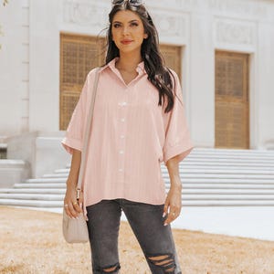 A woman wearing a pink oversized button-down shirt and distressed black jeans, accessorized with a white purse and sunglasses.