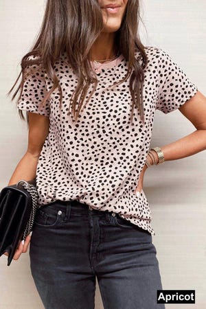 The tee is apricot with a black abstract dot pattern, short sleeves, and a round neckline, paired with dark jeans and accessorized with bracelets and a chain-strap clutch.